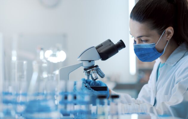Female scientist working in the lab, she is wearing a surgical mask and using a microscope.